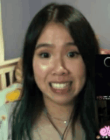 a woman with long black hair is smiling and looking at the camera while sitting in front of a bed .