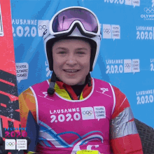 a young boy wearing a helmet and goggles is smiling in front of a blue wall that says lausanne 2020