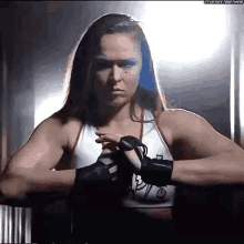 a woman in a white tank top and black gloves is standing in a dark room with her hands on her hips .