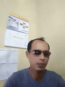 a man wearing sunglasses looks at the camera in front of a wall calendar