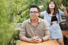 a man wearing glasses sits at a table with his hands folded and a woman standing behind him