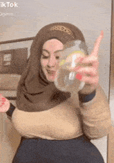 a woman in a hijab is holding a glass of lemonade and pointing at the camera .