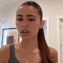 a woman in a tank top and ponytail is standing in a room and looking at the camera .