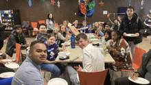 a group of young people are sitting around a table with plates of food and a man wearing a purple shirt that says new life