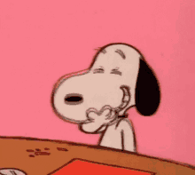 snoopy is sitting at a table and making a heart with his hands .