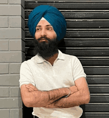 a man with a beard wearing a blue turban and a white shirt is standing with his arms crossed .