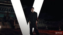 a man in a black coat stands in front of a large letter v and a netflix logo