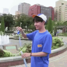 a young man wearing a blue shirt and a baseball cap is standing in front of a fountain holding a straw .