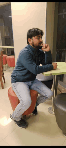 a man in a blue sweater sits on a red stool at a table