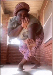 a woman is standing on one leg in front of a brick wall and covering her face with her hands .