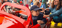 a little boy wearing a blue hat with a volkswagen logo on it sits in a red toy car