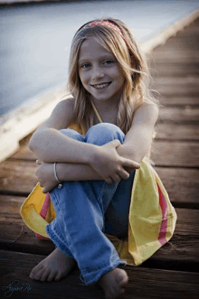 a young girl sits on a wooden dock with her legs crossed and smiles for the camera