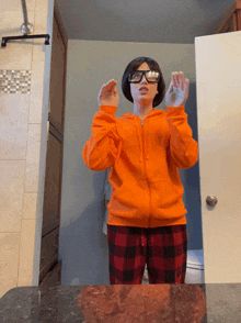 a woman in an orange hoodie and plaid pajamas stands in front of a mirror