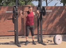 a man in a red tank top is squatting with a barbell on his shoulders in front of a brick wall .