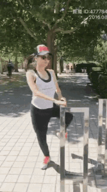 a woman in a white tank top and black leggings is standing on a metal railing on a sidewalk ..