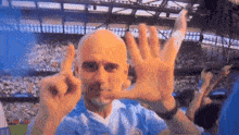 a bald man in a blue shirt holds up his hands in front of a crowd at a soccer game