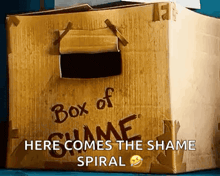 a cardboard box with a hole in it that says `` box of shame here comes the shame spiral '' .