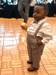 a little boy in suspenders and a bow tie holds a bread roll