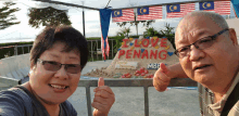 a man and a woman are giving a thumbs up in front of a sand sculpture that says i love penang