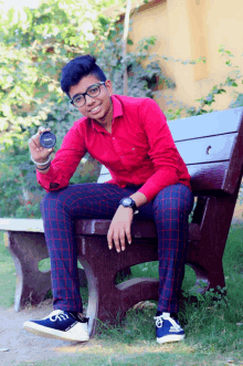 a young man in a red shirt and plaid pants sits on a bench holding a canon camera