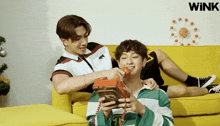 two young men are sitting on a yellow couch and one of them is holding a red phone with the word wink on the bottom