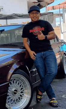 a man in a hard rock cafe t-shirt is standing next to a car and giving a thumbs up .