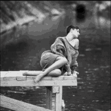 a black and white photo of a woman in a sweater sitting on a wooden dock .