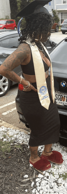 a woman in a graduation cap and gown stands in front of a bmw with a license plate that says sgd11