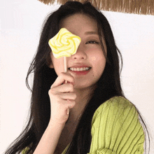 a woman in a green sweater holds a yellow lollipop in front of her eye