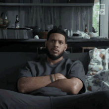 a man with a beard is sitting on a couch with his arms crossed in front of a messy kitchen