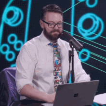 a man wearing glasses and a tie is sitting in front of a microphone with a laptop in front of him