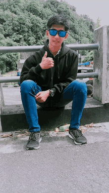 a young man wearing sunglasses and a black hoodie sits on a ledge and gives a thumbs up