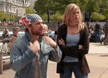 a man wearing an american flag bandana and a denim jacket is standing next to a woman with her arms crossed .