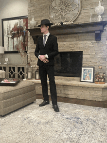 a man in a black suit and hat stands in front of a fireplace