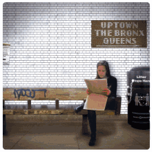 a woman sits on a bench reading a newspaper in front of a sign that says uptown the bronx queens on it