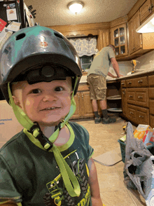 a little boy wearing a helmet and a john deere shirt smiles in a kitchen