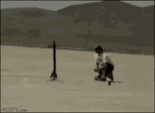 a man is kneeling on the ground in the desert while another man takes a picture of a plane flying overhead .