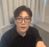 a young man wearing glasses and a black shirt is sitting at a table in a room .