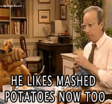 a man in a suit and tie is sitting at a table eating mashed potatoes with alf .