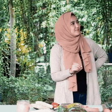 a woman wearing a hijab and a cardigan is standing in front of a table with food on it .