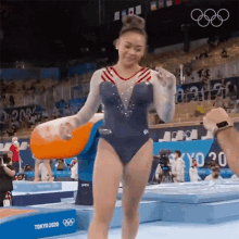 a female gymnast in a blue leotard is standing on a mat in front of a crowd at the olympics .