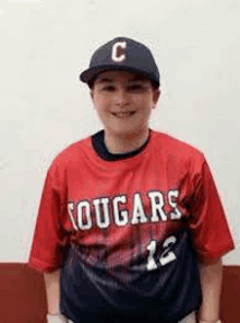 a young boy wearing a cougars baseball jersey and hat is standing in front of a wall .