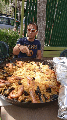 a man wearing a shirt that says ' rs ' on it sits at a table with a pan of food