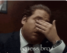 a man in a suit and tie covering his face with his hand and the words " i guess bro " behind him