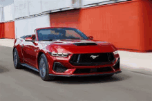 a red ford mustang convertible is driving down a street in front of a red wall .