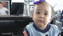 a baby girl with a purple bow on her head is making a funny face while sitting in front of a desk .