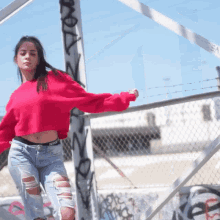 a woman wearing a red crop top and ripped jeans is standing in front of a fence with graffiti on it