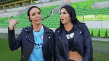 two women are standing next to each other on a soccer field . one of the women is wearing a manchester city jersey .