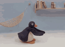 a stuffed penguin is squatting on the floor in front of a hammock and a bookshelf .