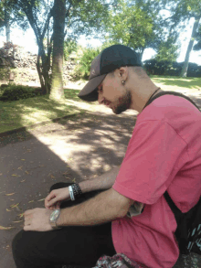 a man wearing a pink shirt and a black hat is sitting on the ground looking at his watch